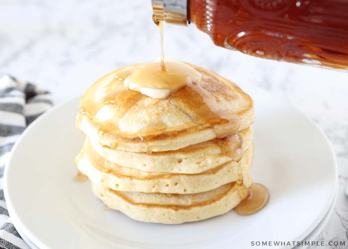 pouring syrup on top of a stack of pancakes with a slab of butter